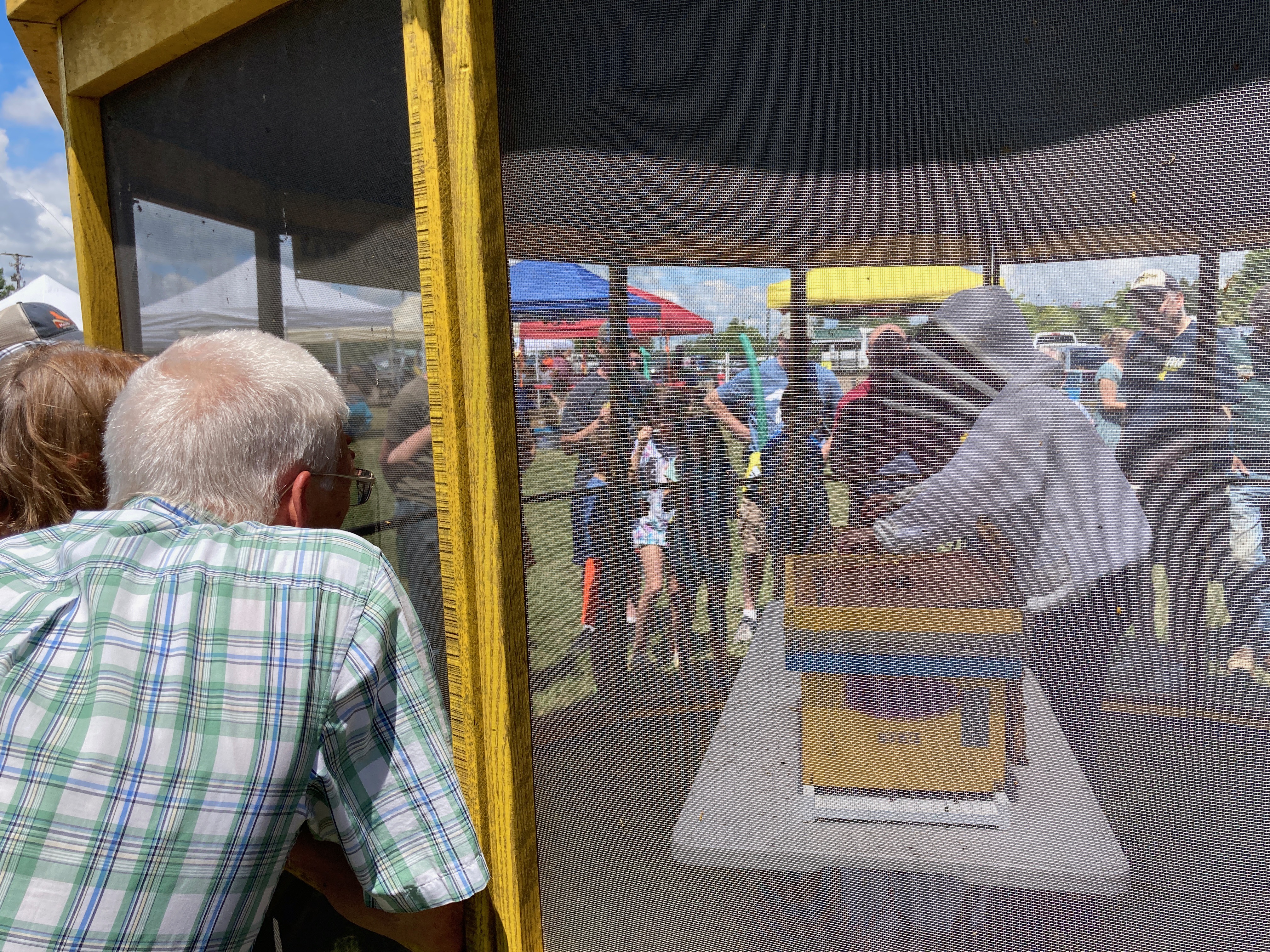 Photo of people gathered around a screened tent and a beekeeper in the tent